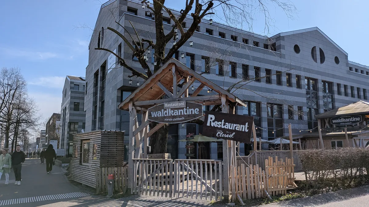 Blick auf ein Gartenrestaurant vor einem dunkel-hell gestreiften Bürogebäude.